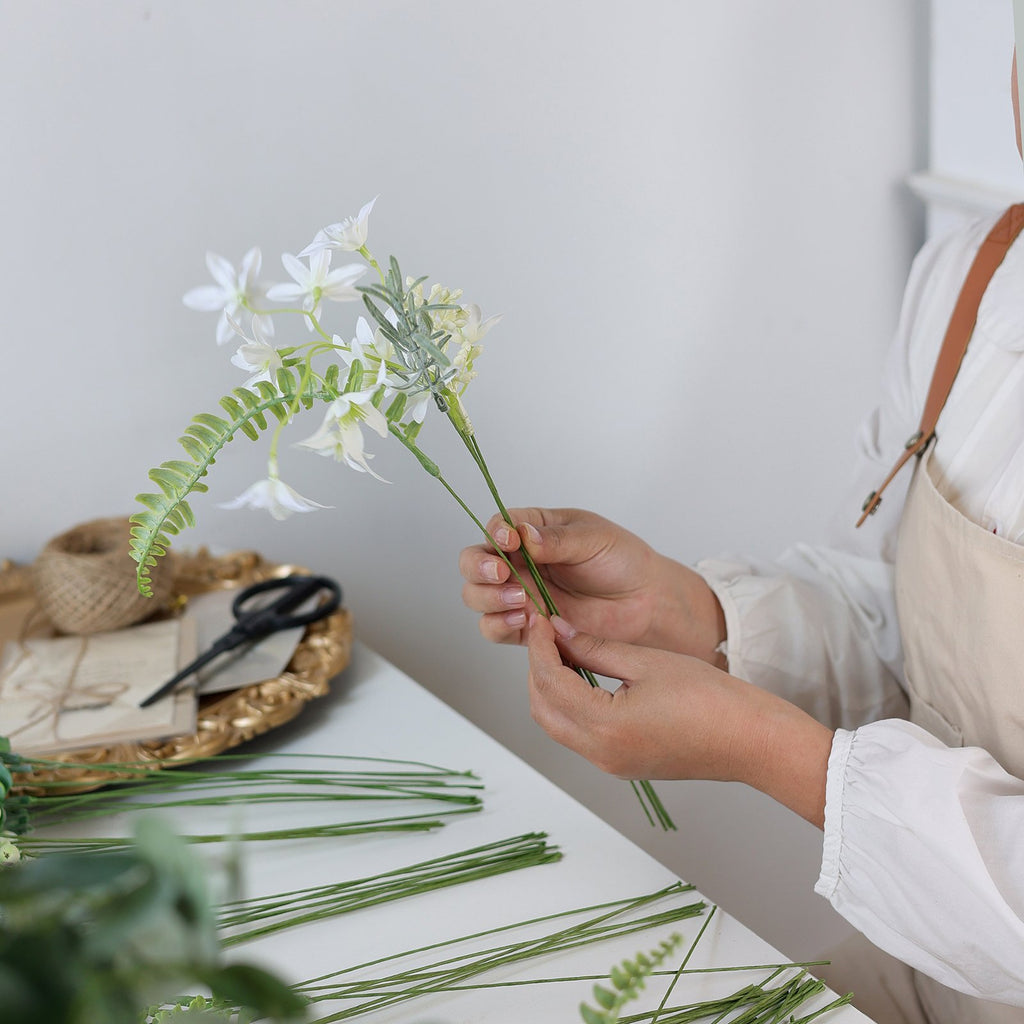 Hands assembling artificial greenery stems with white flowers and fern leaves, with crafting supplies on a tray.