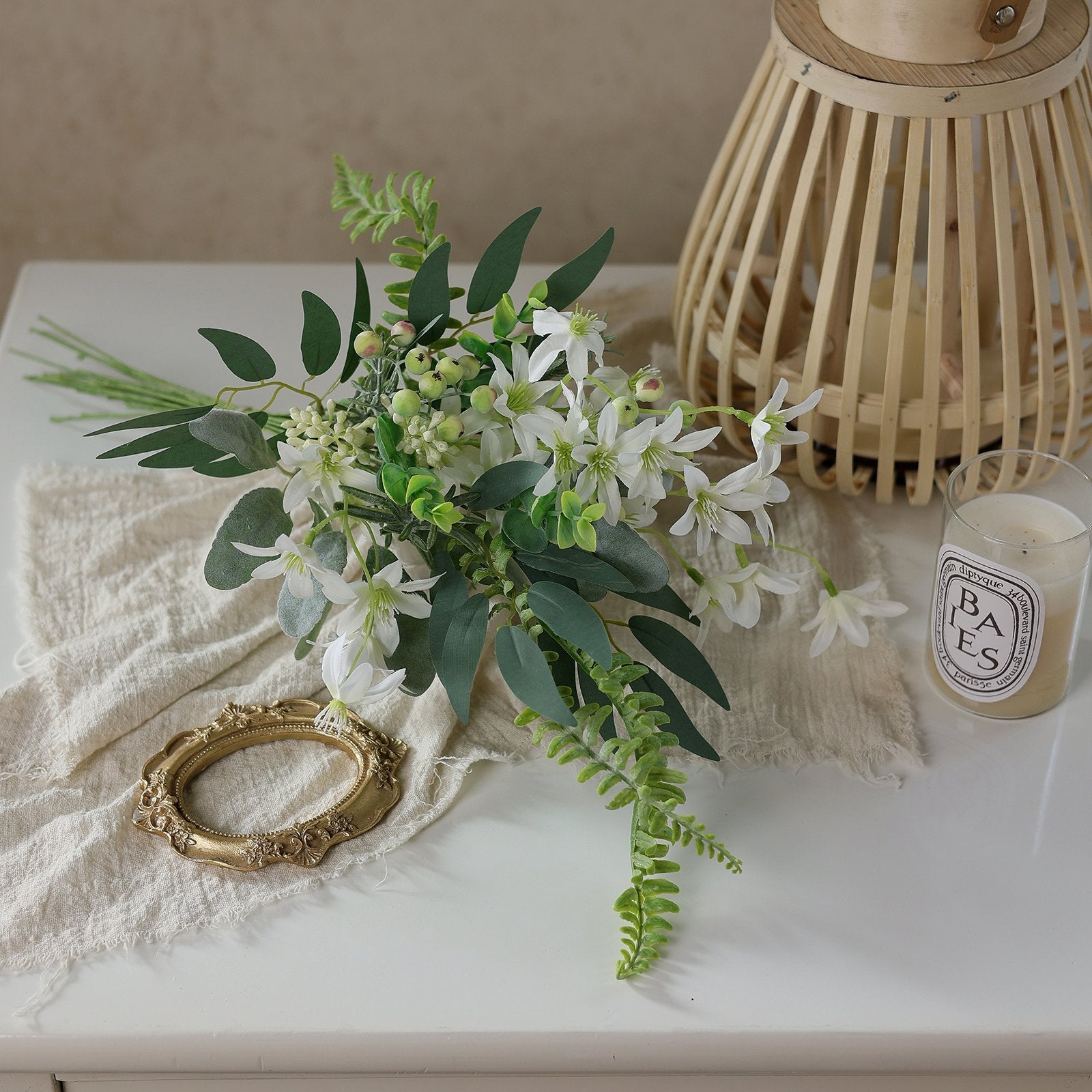 Artificial greenery bouquet with white flowers, berries, and ferns on a textured cloth next to a decorative lantern and candle.