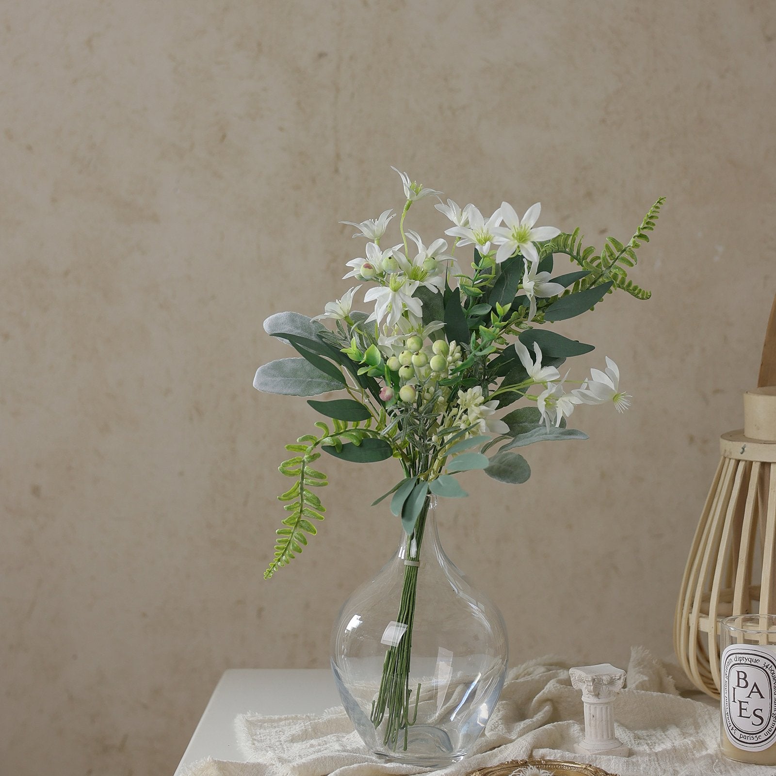Bouquet of artificial white flowers and green foliage in a clear glass vase on a white cloth.