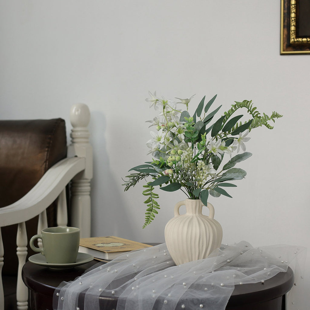 Artificial greenery stems in a cream vase on a table with a book and cup