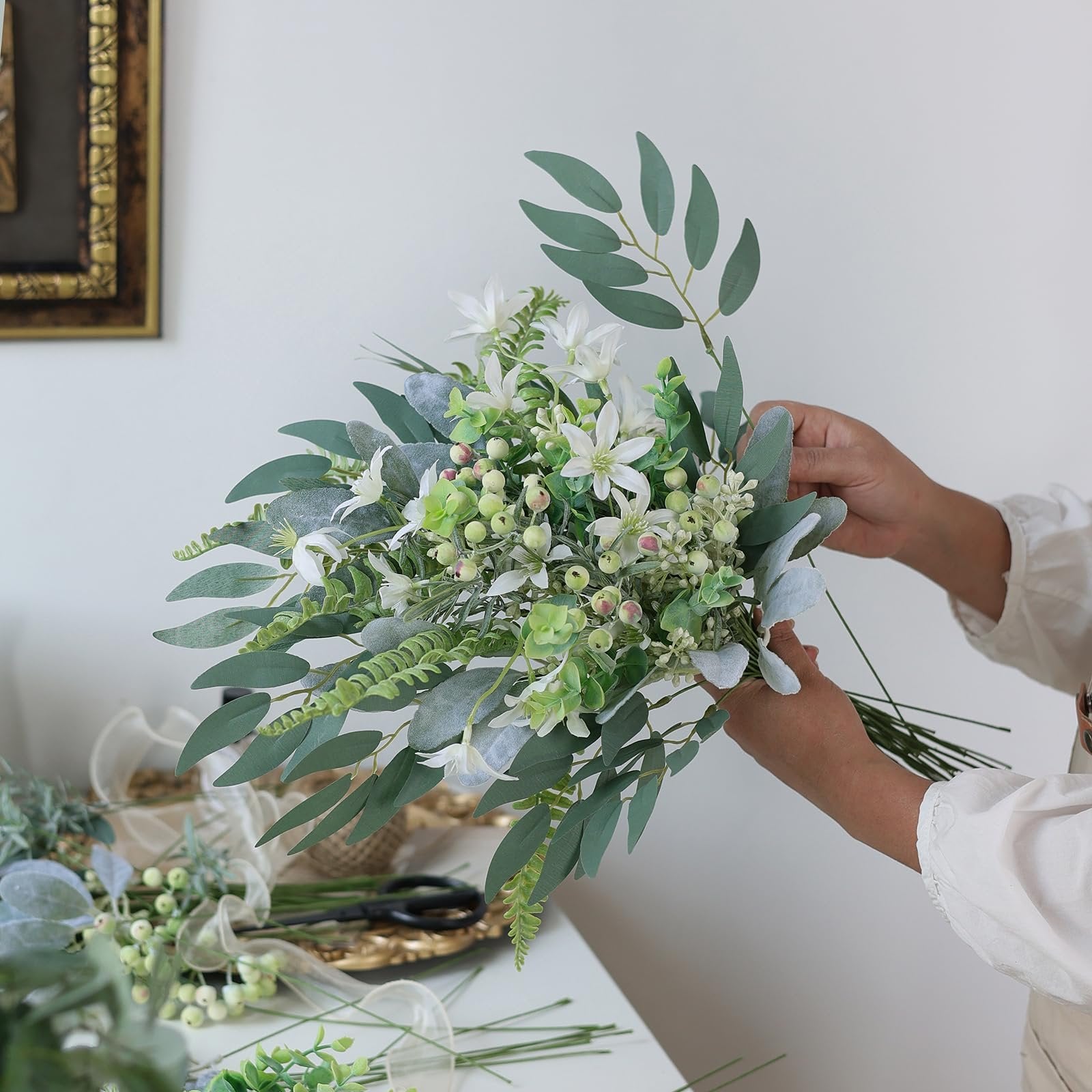 Hands arranging a bouquet of artificial greenery stems including eucalyptus, fern, and berries.