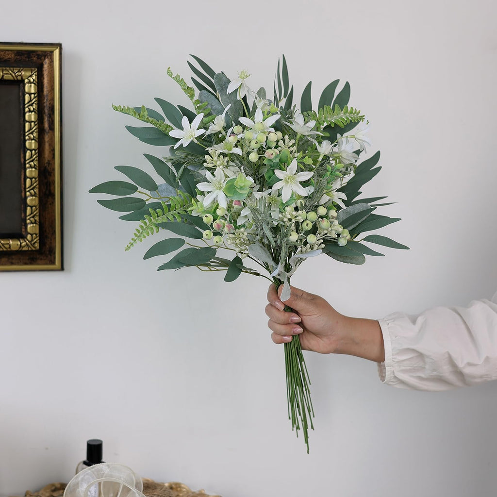 Hand holding a bouquet of artificial greenery stems with white flowers and berries against a white wall.