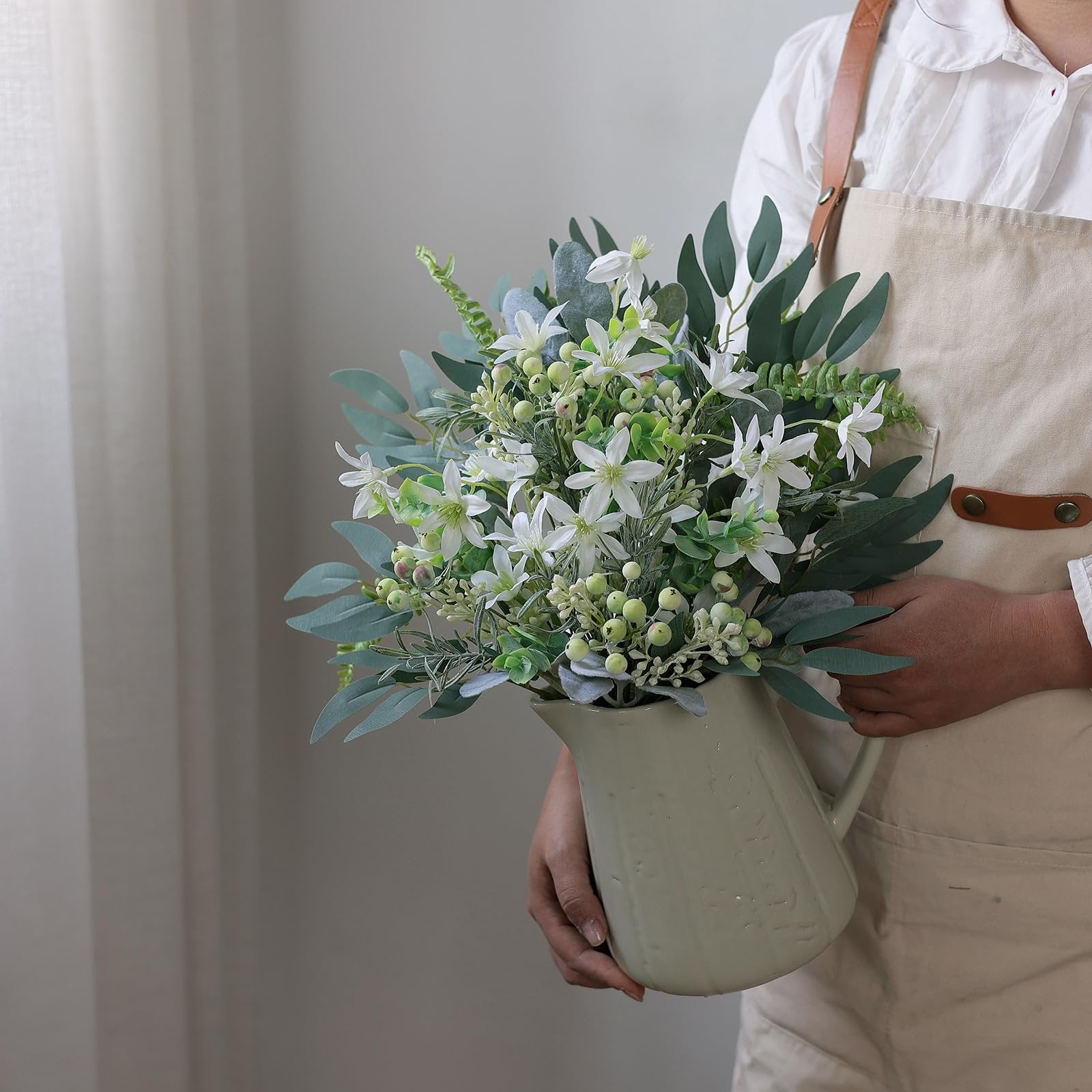 Person holding a light green pitcher filled with artificial greenery and white flowers, wearing an apron.