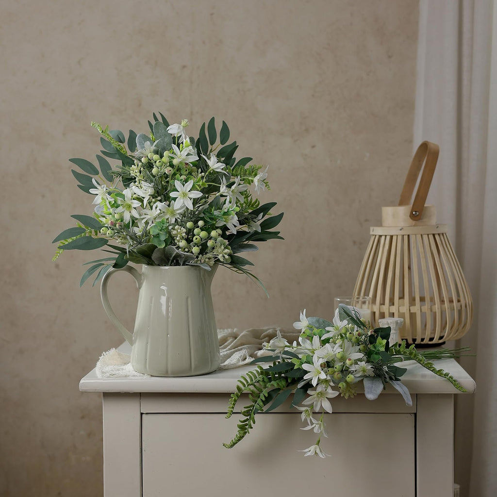 Two bouquets of artificial white flowers and greenery arranged in a cream pitcher and on a table, with a wooden lantern in the background.