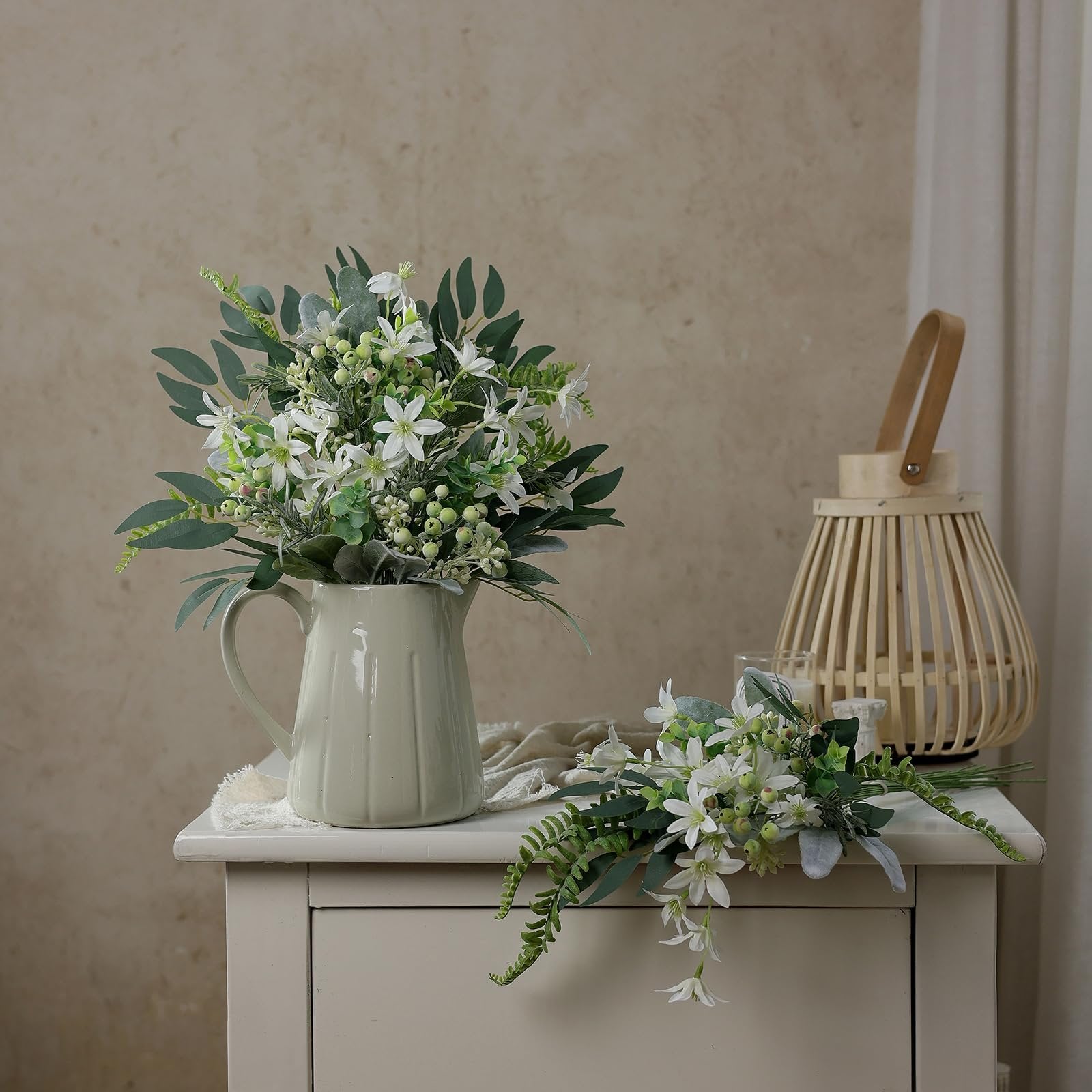 Two bouquets of artificial white flowers and greenery arranged in a cream pitcher and on a table, with a wooden lantern in the background.