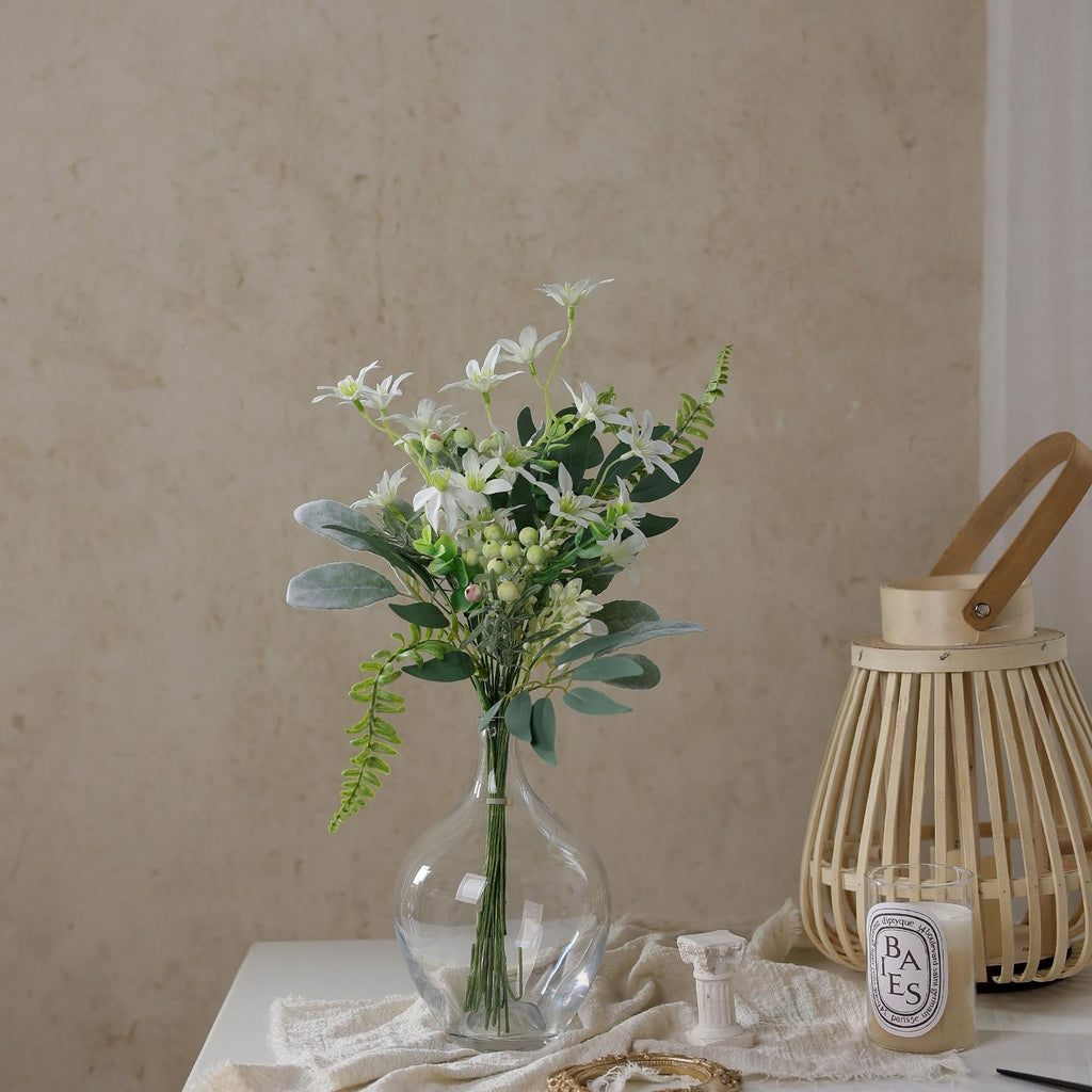 Artificial greenery stems with white flowers in a clear glass vase on a table with a candle lantern.