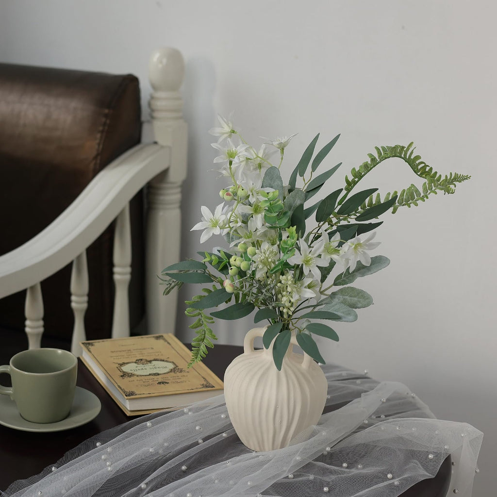 Artificial greenery stems with white flowers in a vase on a table next to a book and mug.