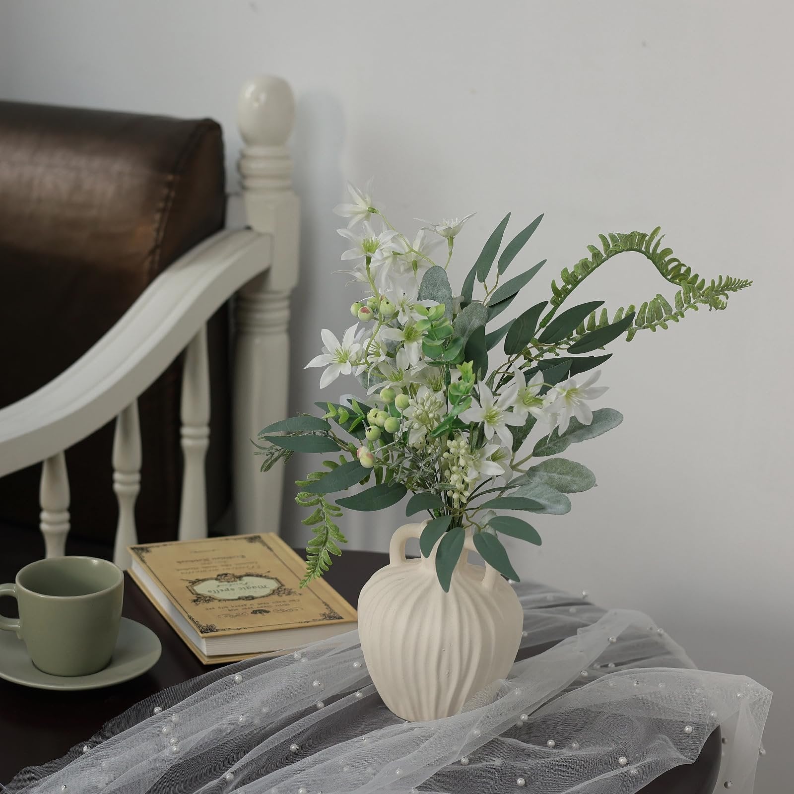 Artificial greenery stems with white flowers in a vase on a table next to a book and mug.