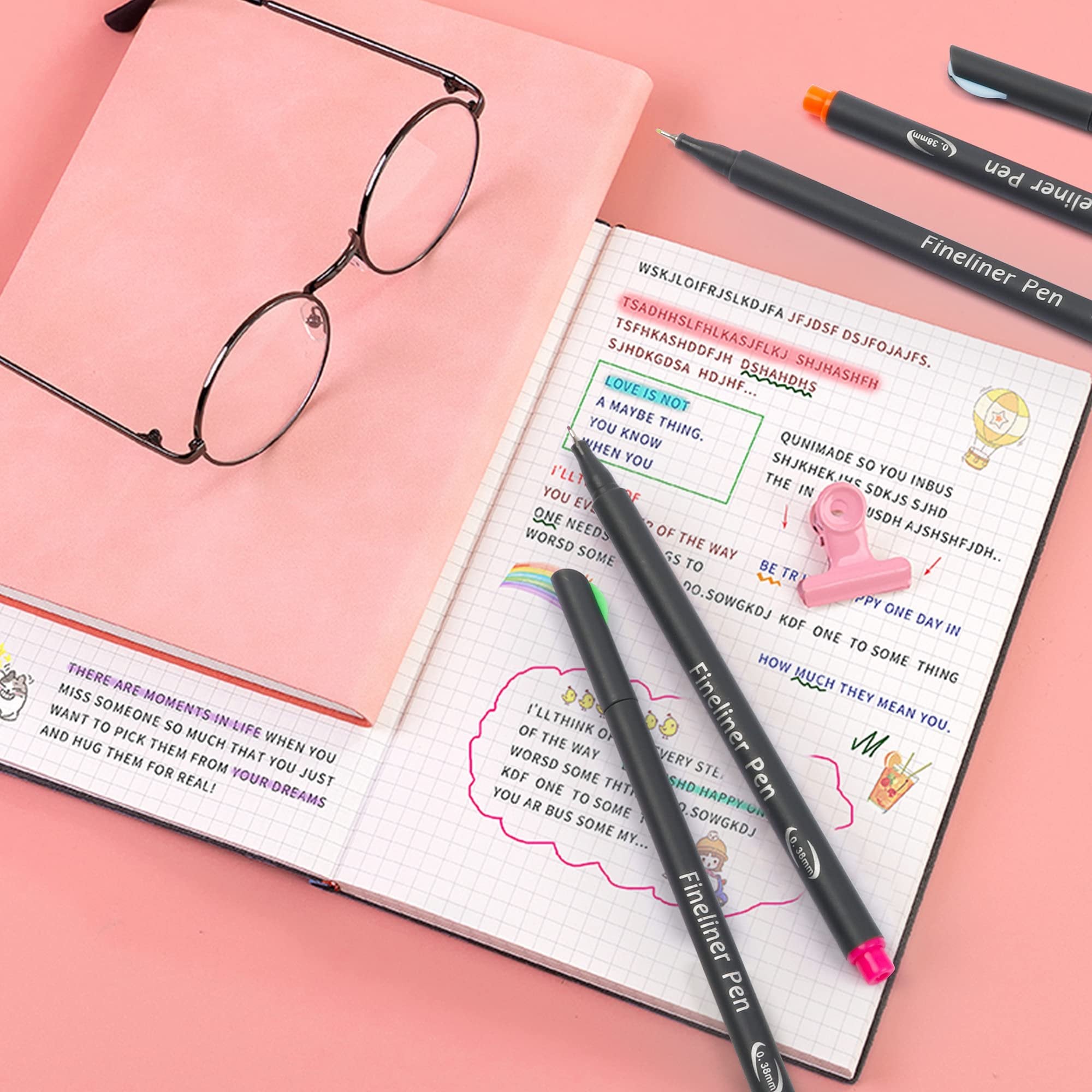 Fineliner pens laid out on an open notebook with glasses and a pink background.