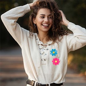 Woman with curly hair wearing a cream sweater with two embroidered flower patches, one blue and one pink, on the chest.