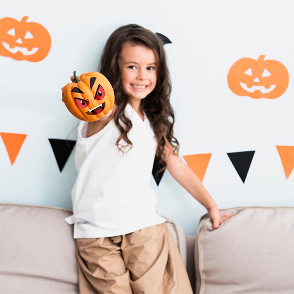 Smiling girl holding a decorated pumpkin with a spooky face, with Halloween decorations in the background.
