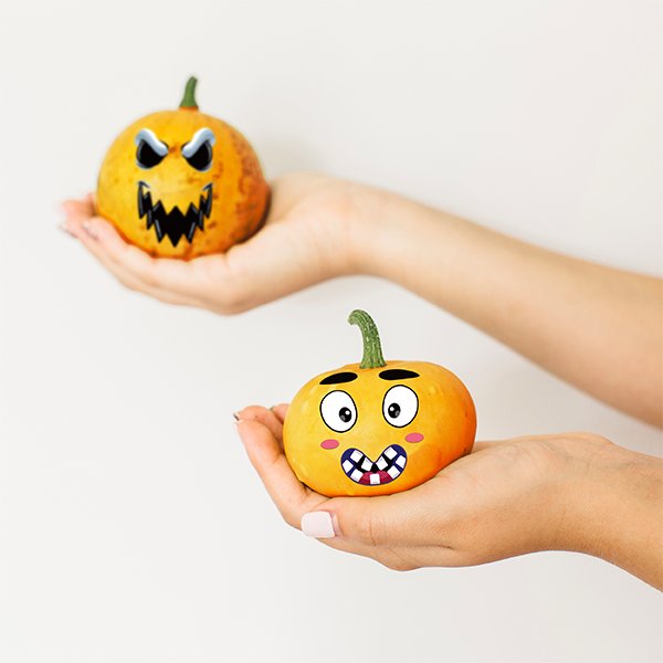 Two small pumpkins decorated with Halloween face stickers, held in hands against a white background.