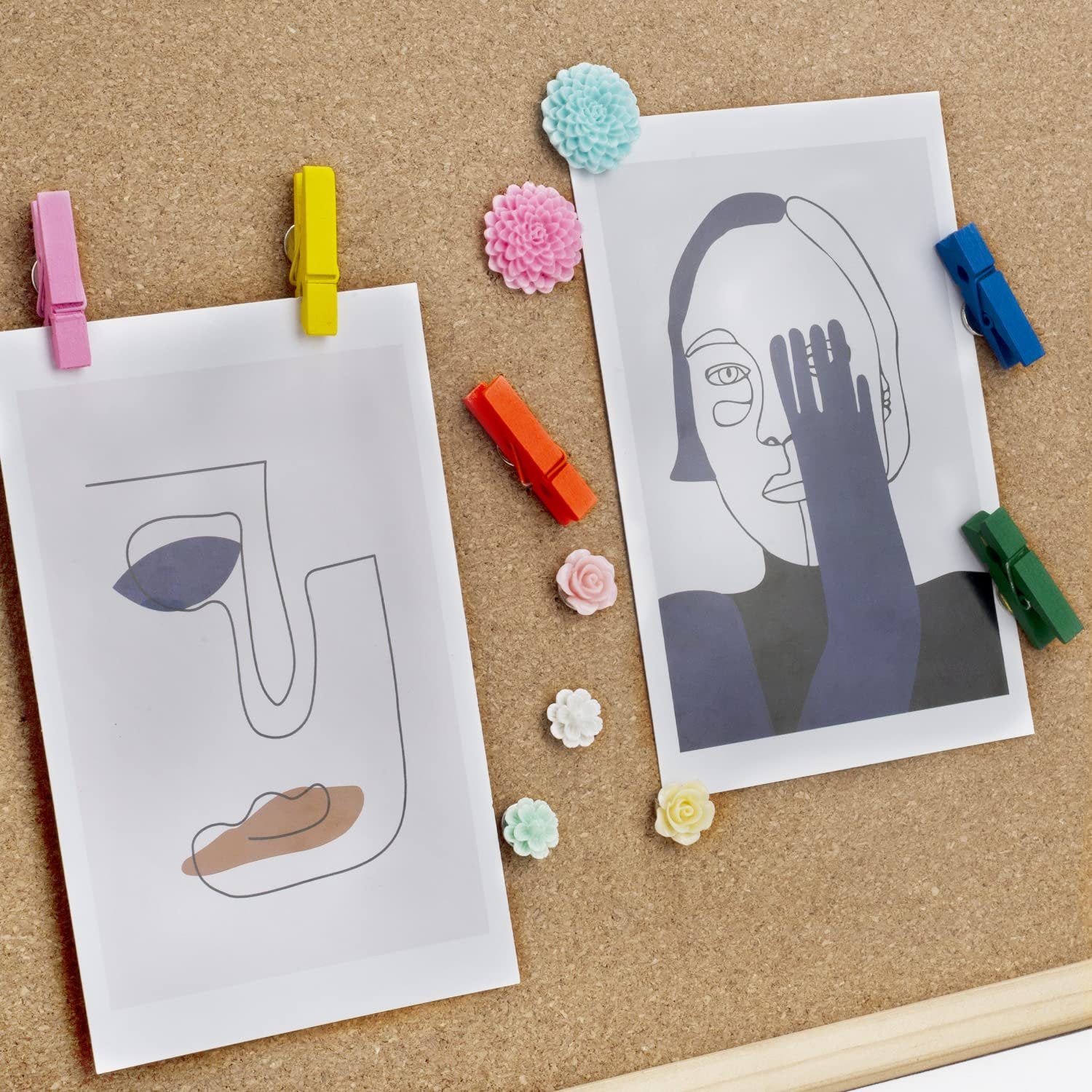Corkboard with two abstract art prints pinned by colorful clothespins and decorative flower-shaped push pins.
