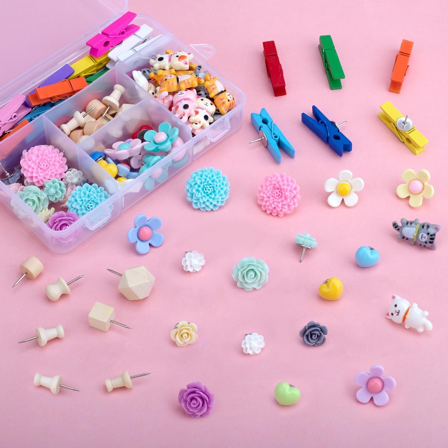 Assorted decorative push pins and mini clothespins in a clear organizer box and scattered on a pink background.
