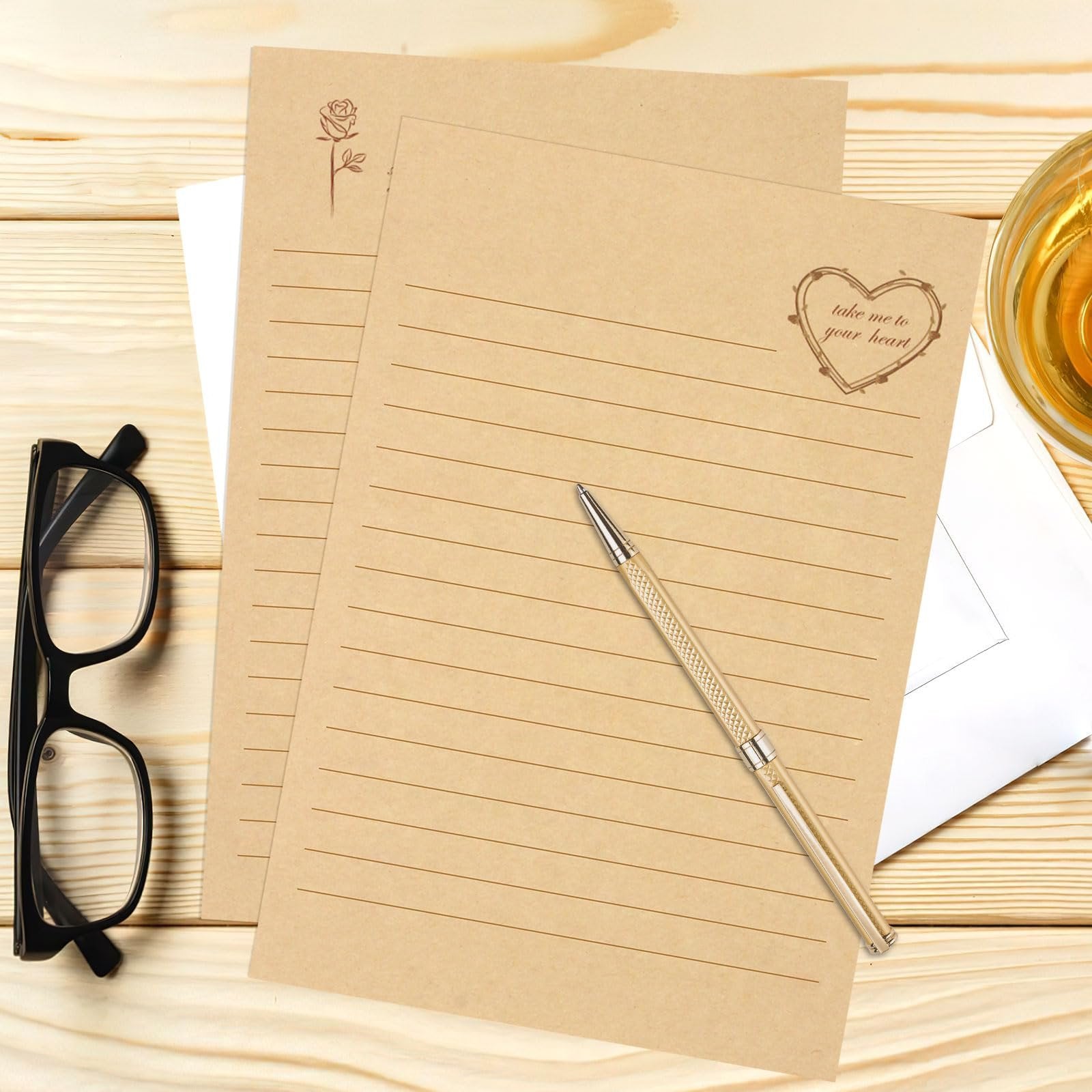 Vintage stationery set with lined kraft paper, pen, glasses, and a glass of amber liquid on a wooden desk.