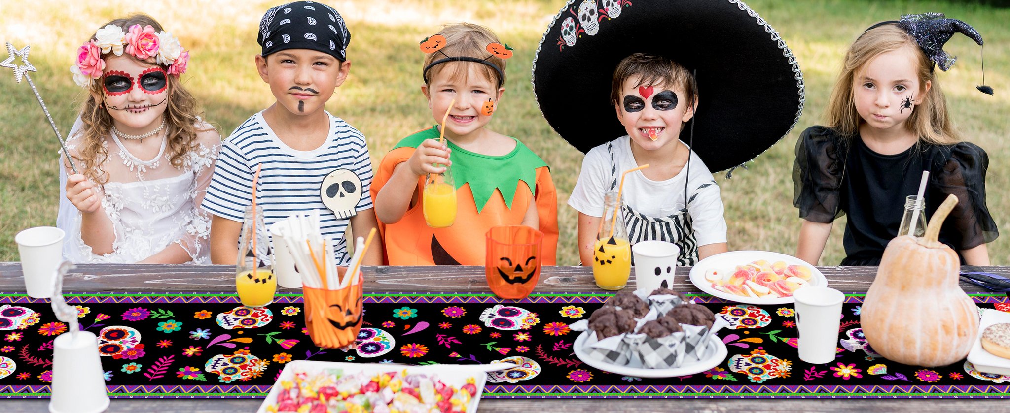 Five children in Halloween costumes sit at a picnic table with a sugar skull print table runner.