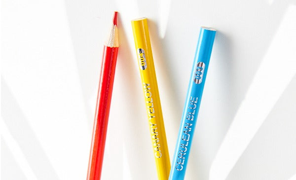 Three colored pencils, red, yellow, and blue, on a white background with logo in the upper right corner.