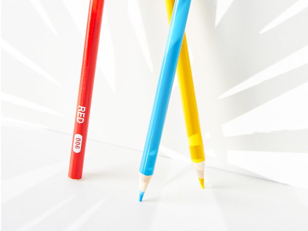 Three colored pencils, red, blue, and yellow, angled against a white background with shadow patterns.