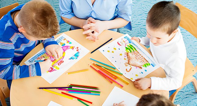 Children drawing with colored pencils at a table with an adult supervising