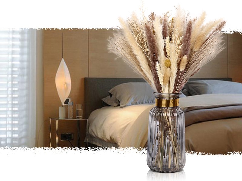Dried pampas grass arrangement in a ribbed glass vase on a bedside table with a bed in the background.