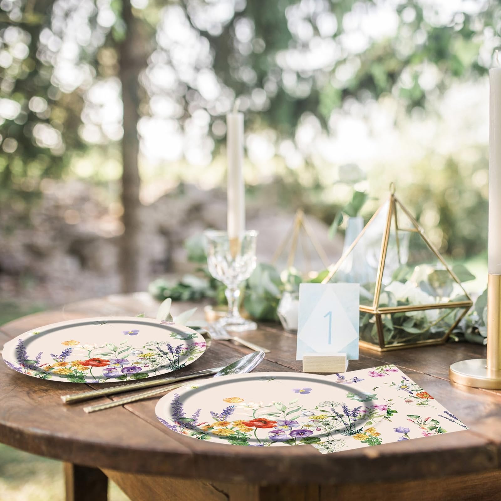 Floral paper plates and napkins set on a wooden table with candles and greenery, featuring a table number '1'.