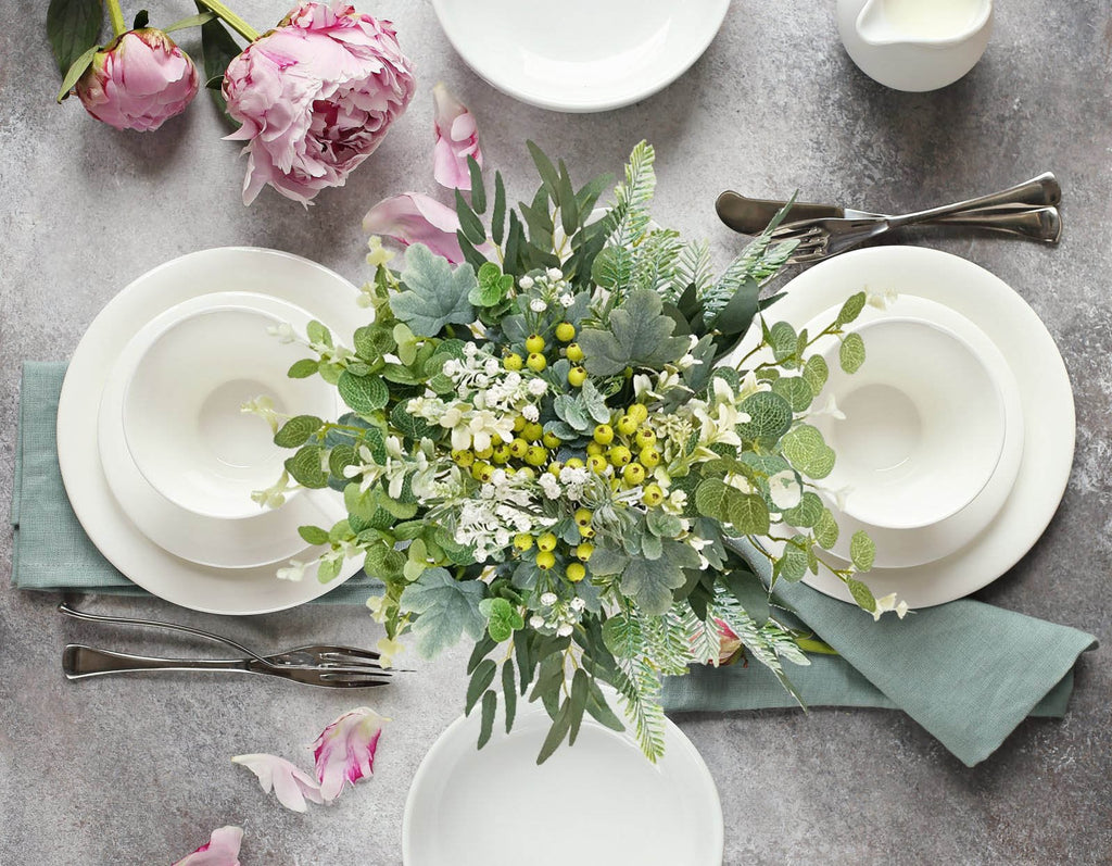 Table setting with a floral arrangement of artificial greenery, white plates, bowls, and cutlery, accented by pink peonies.