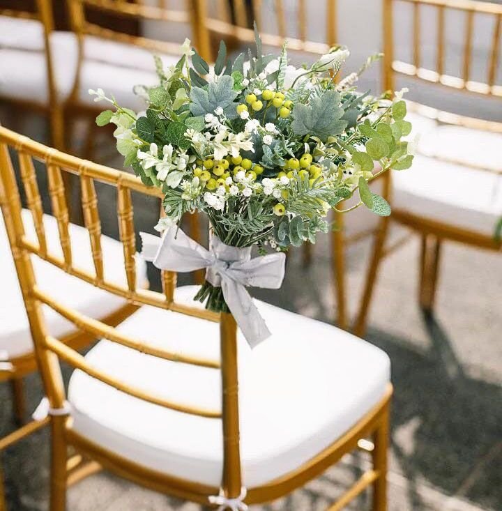 Artificial greenery bouquet with yellow berries and white flowers tied with a gray ribbon, resting on a gold chair with a white cushion.