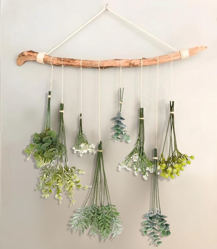 Hanging wall decor with a driftwood branch and assorted artificial greenery stems suspended by twine.