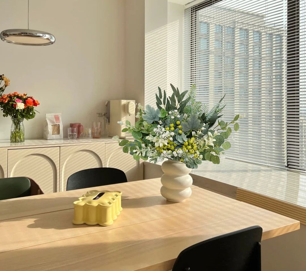 Artificial greenery stems in a white vase on a wooden table, with blinds in the background.