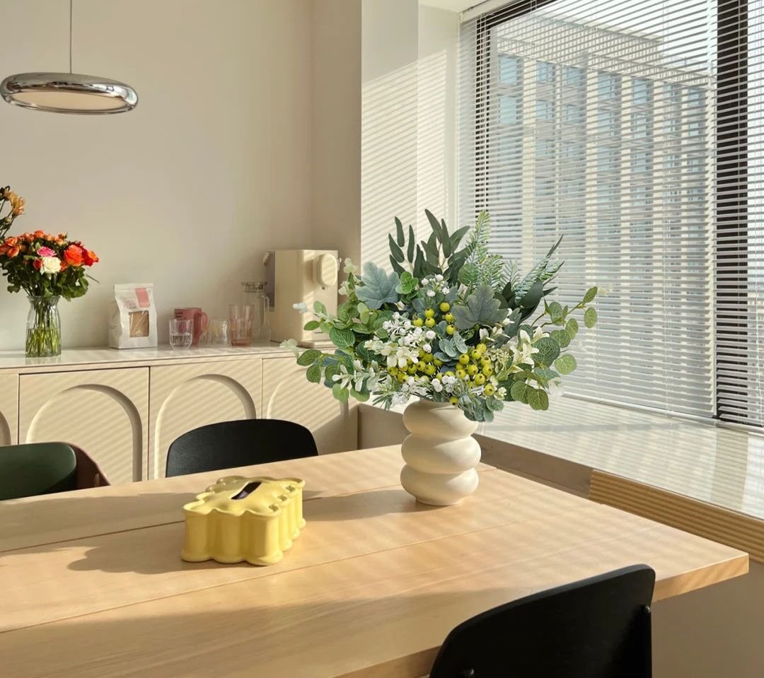 Artificial greenery stems in a white vase on a wooden table, with blinds in the background.