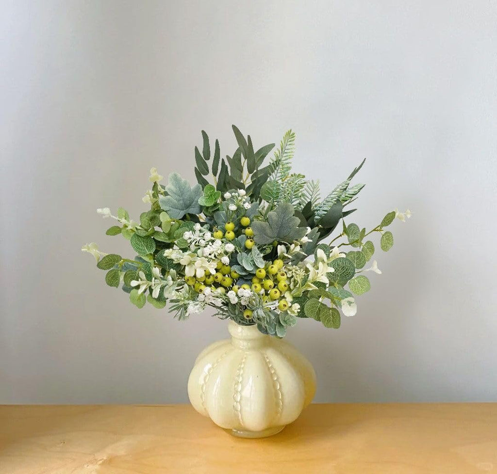 Artificial greenery stems with white and yellow accents in a cream vase on a wooden surface.