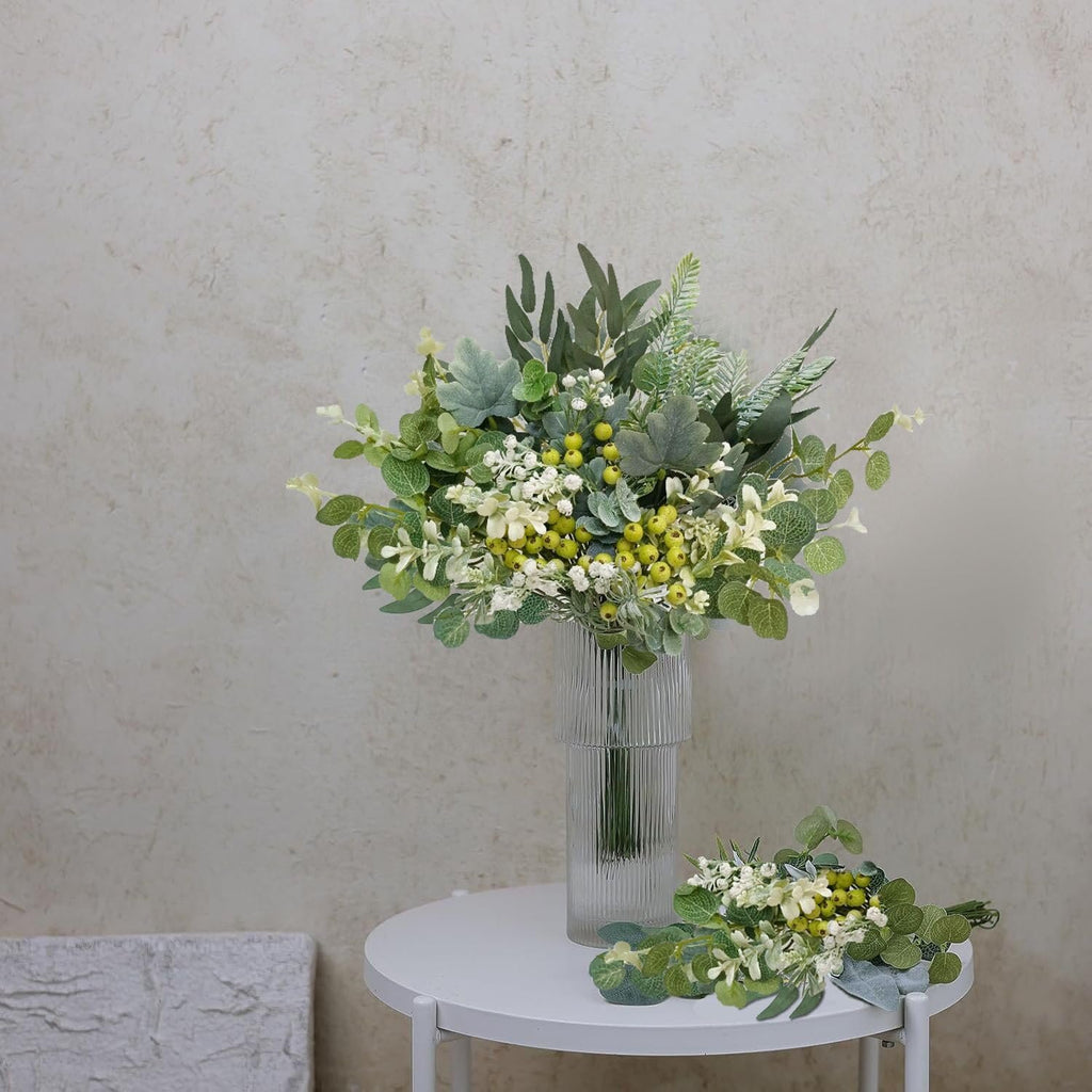 Artificial greenery stems arranged in a vase and on a table, featuring green leaves and yellow berries.