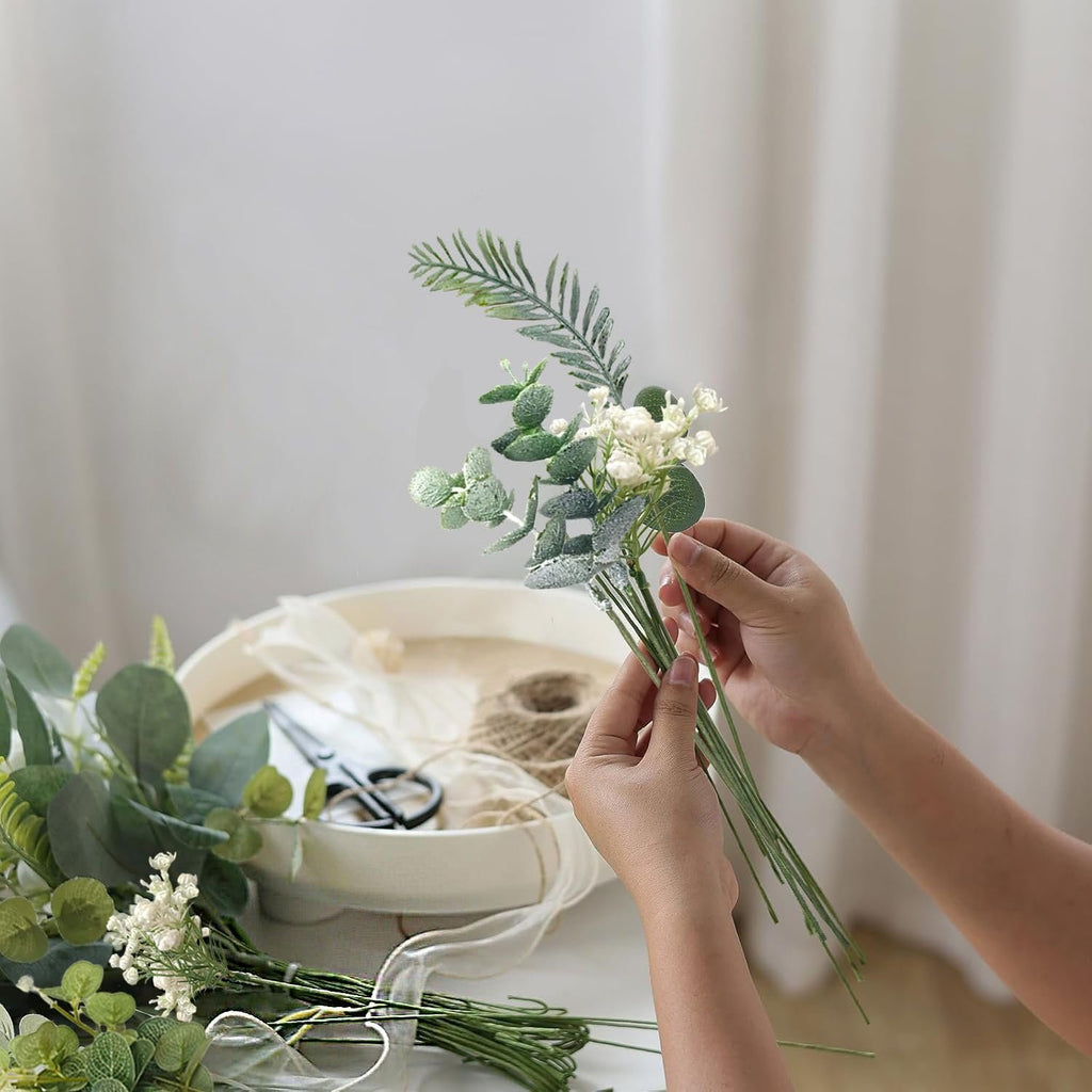 Hands arranging artificial greenery stems and small white flowers for a floral arrangement.