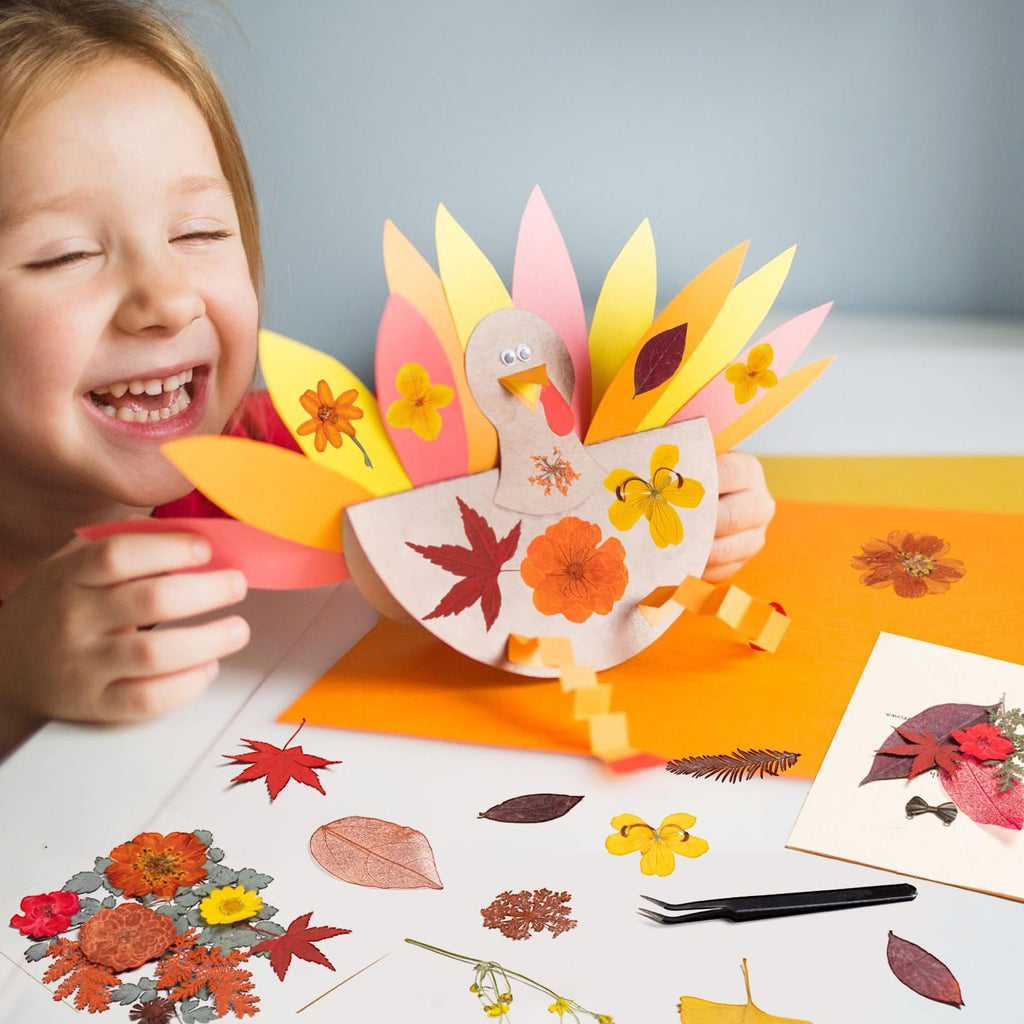 Young girl laughing while holding a paper turkey craft decorated with dried fall flowers and leaves. Various dried botanicals and tweezers are scattered on the table.