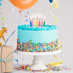 Light blue frosted birthday cake with colorful sprinkles and lit candles on a white cake stand, with a dinosaur toy and gift box.