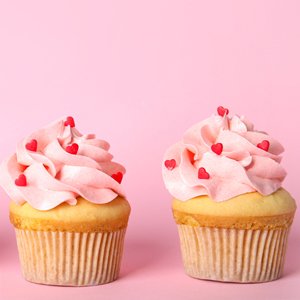 Two frosted cupcakes with pink frosting and red heart sprinkles on a pink background