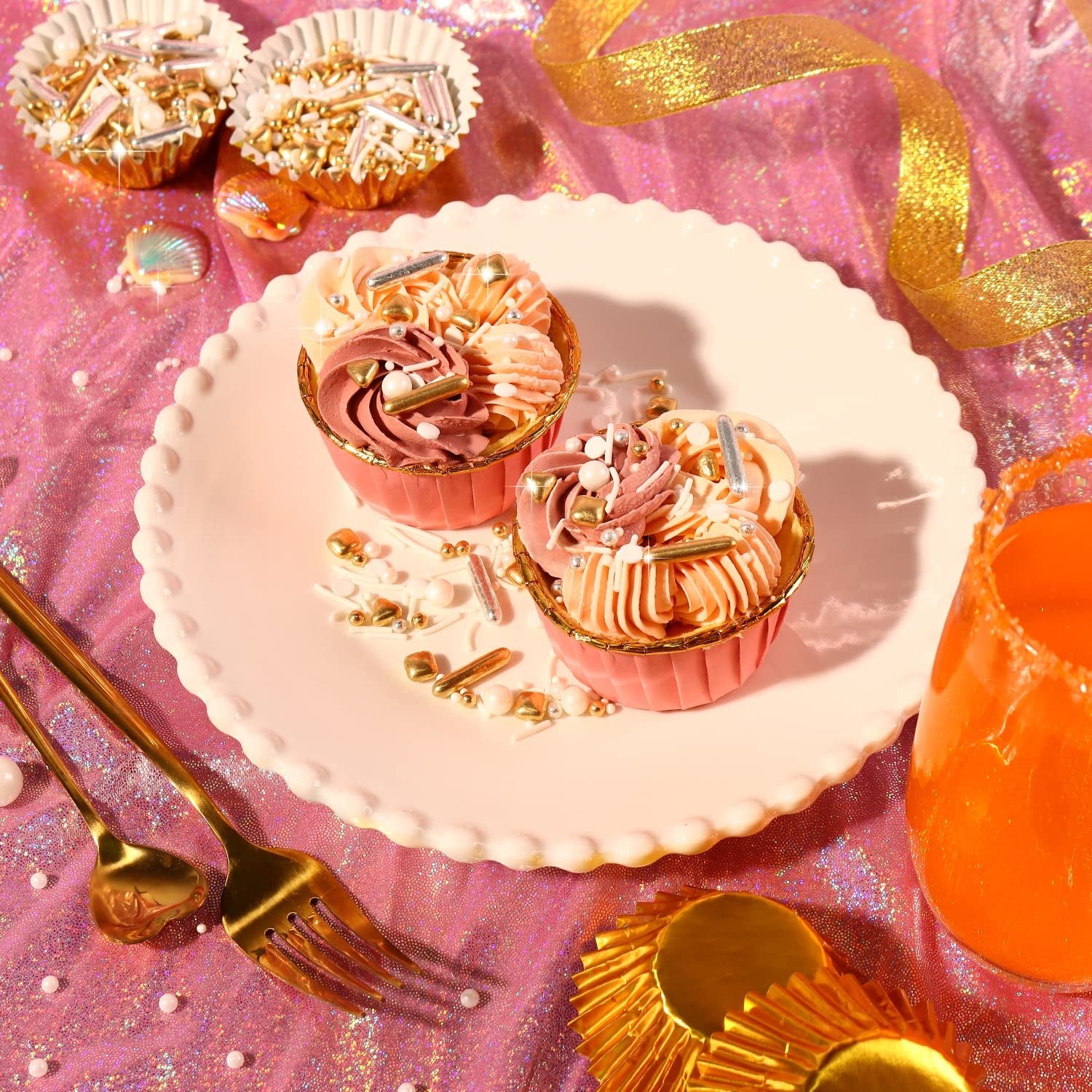 Two decorated cupcakes with gold and silver sprinkles on a white plate, surrounded by sprinkles and gold ribbon.