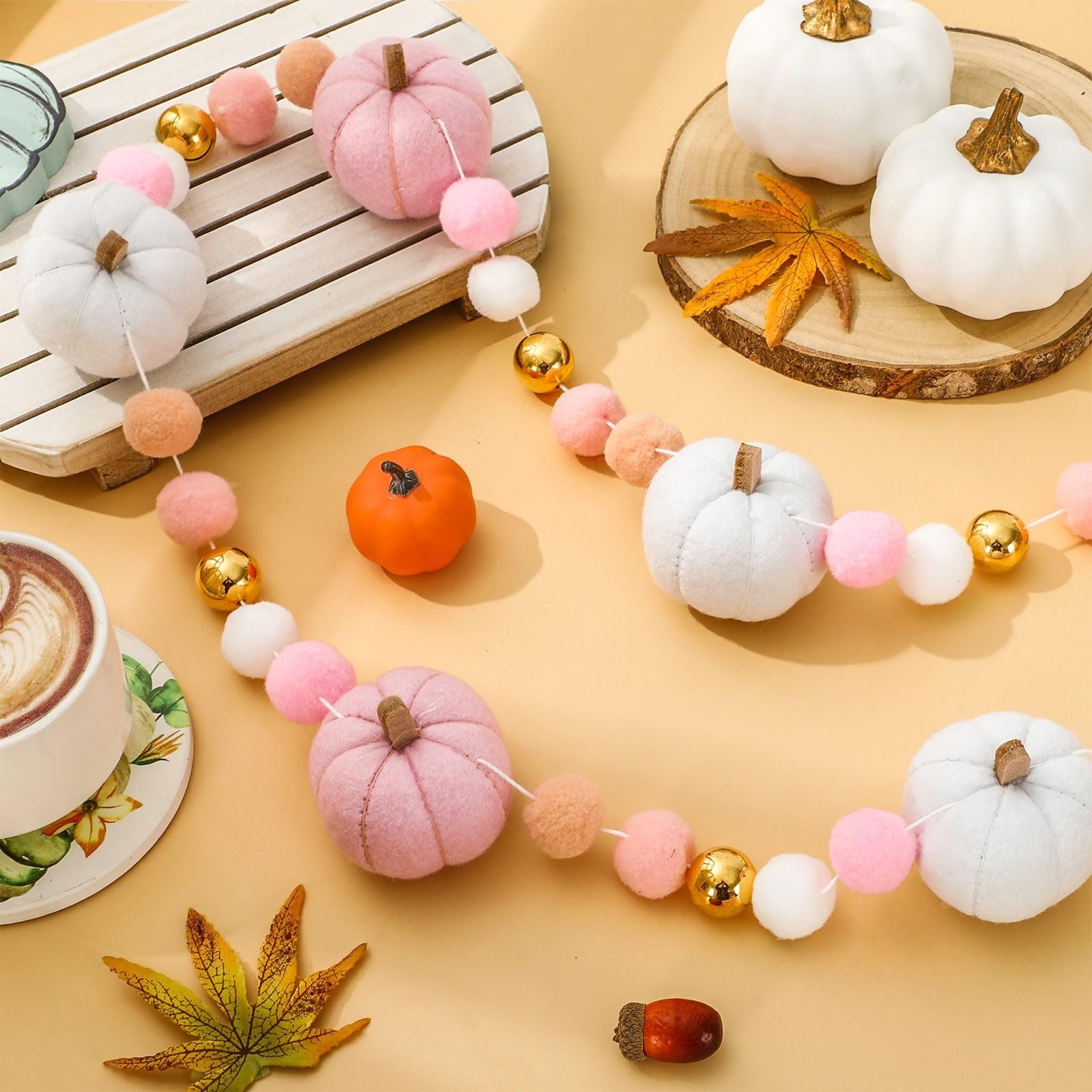 Fall felt garland with pink, white, and orange pumpkins, pom poms, and gold beads, arranged with a coffee cup and autumn leaves.