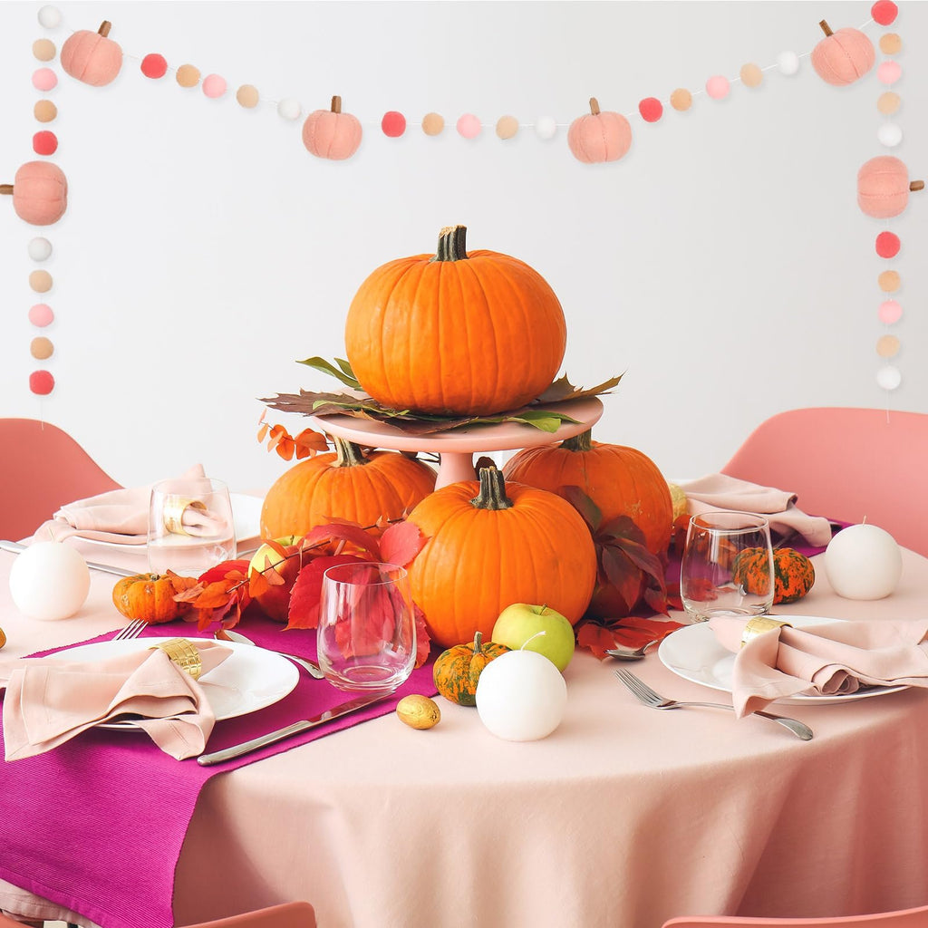 Fall table setting with pumpkins, felt ball garland, and autumn leaves on a pink tablecloth.