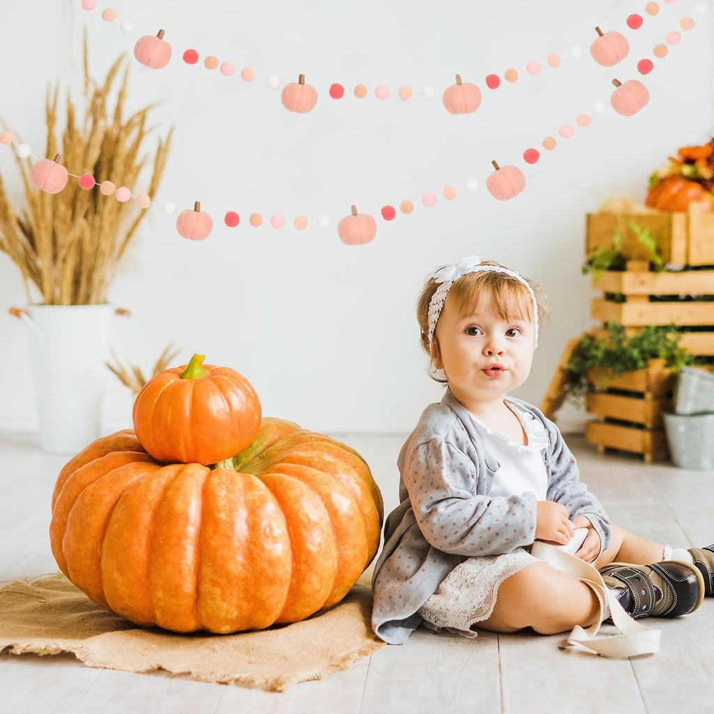 Baby sitting next to a large pumpkin with a felt ball garland featuring pumpkins hanging above