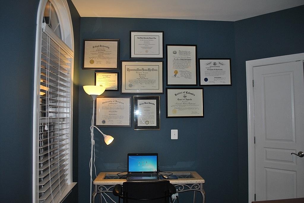 Wall of framed certificates and diplomas above a desk with a laptop and lamp.