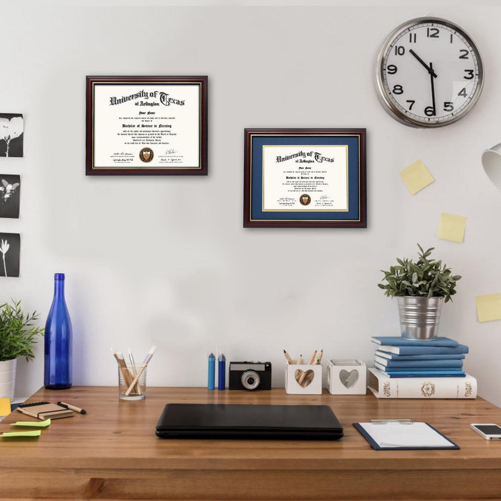 Two framed diplomas on a wall above a wooden desk with office supplies and a clock.