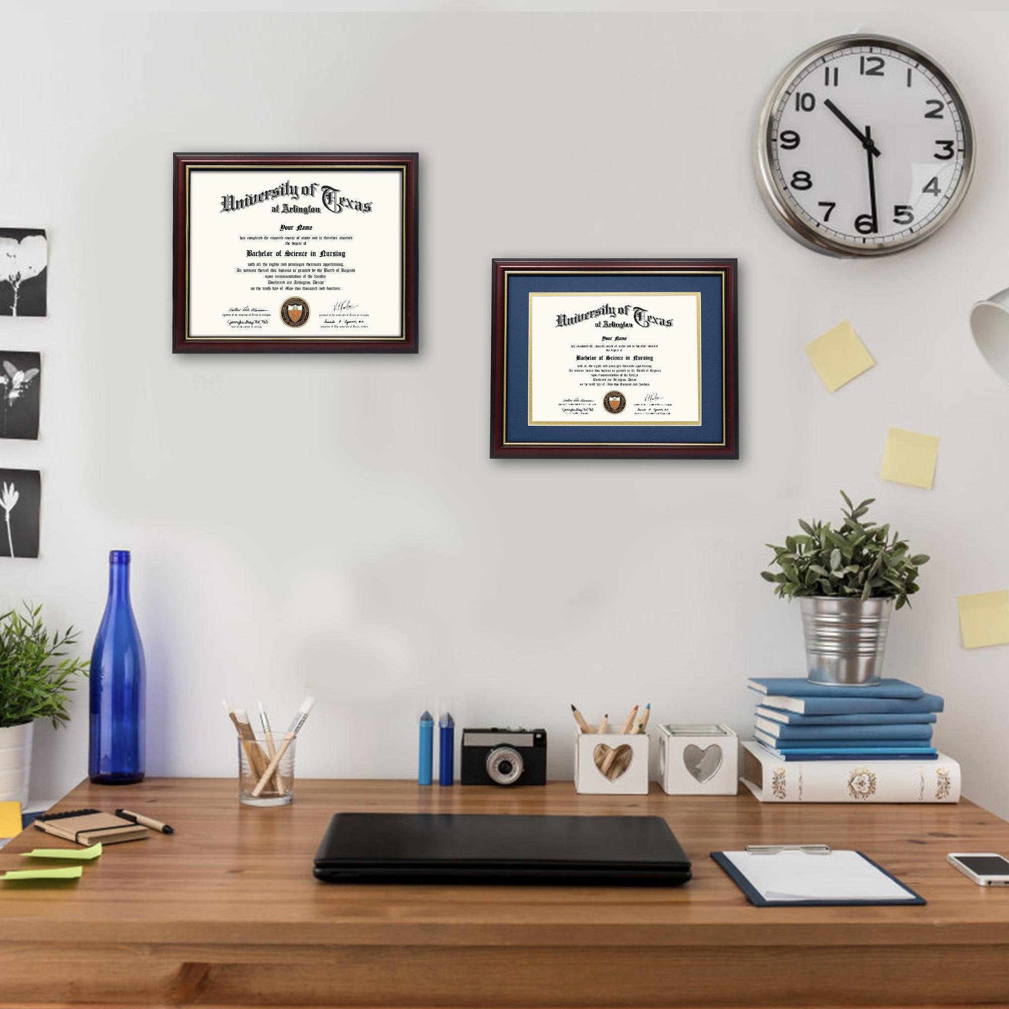 Two framed diplomas on a wall above a wooden desk with office supplies and a clock.