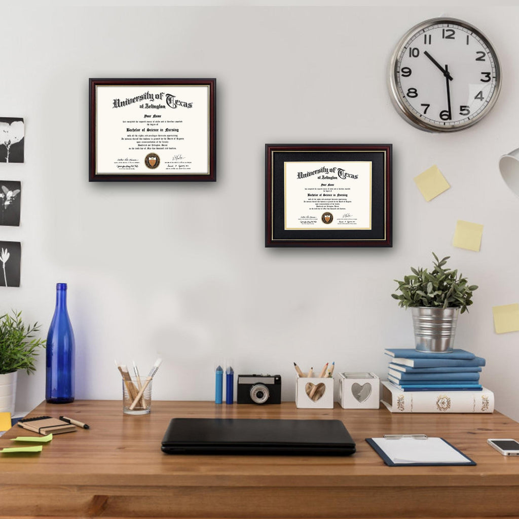 Two framed diplomas on a wall above a wooden desk with office supplies and a clock.