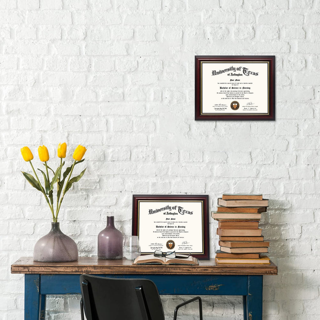 Two diploma frames on a desk with books and flowers, one frame is larger and higher up on the wall.