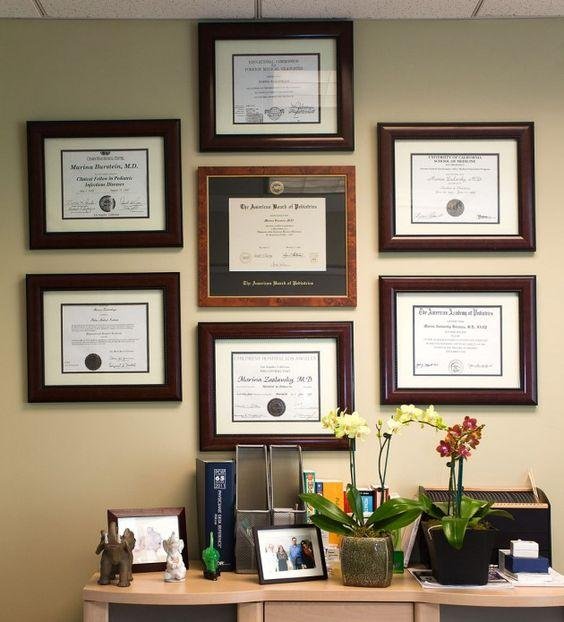 Seven framed diplomas and certificates hanging on a wall above a desk with plants and photos.
