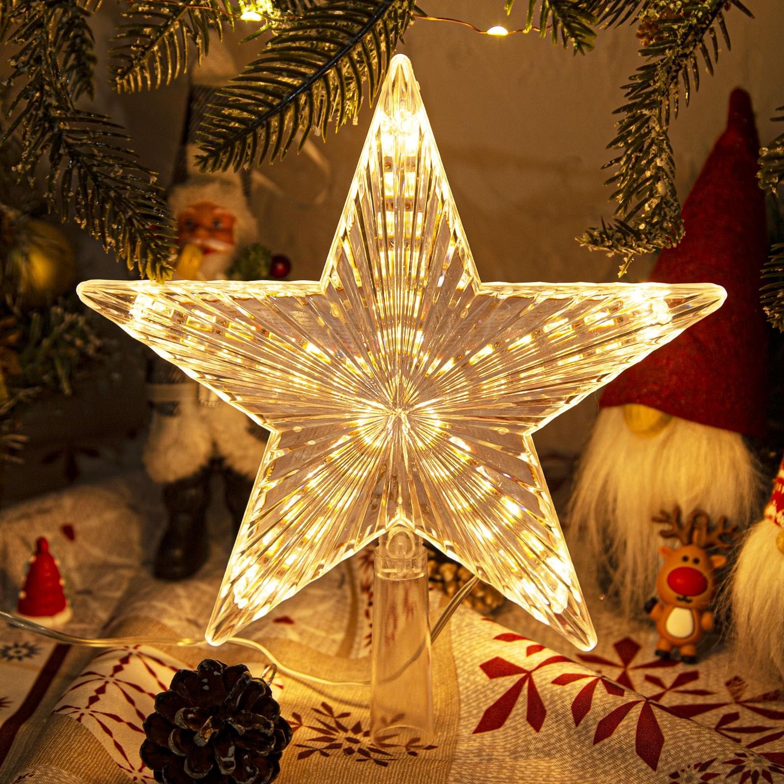 Illuminated star tree topper with warm white LED lights, featuring a ribbed prismatic design, set against a festive background with pine branches and decorative gnomes.