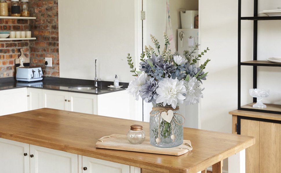 Artificial dahlia flower arrangement in a textured glass vase on a wooden tray, set on a kitchen island.