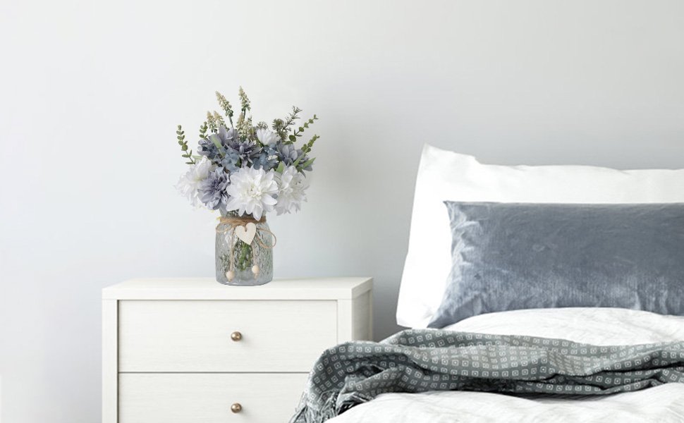 Artificial dahlia flower arrangement in a glass vase on a nightstand next to a bed.