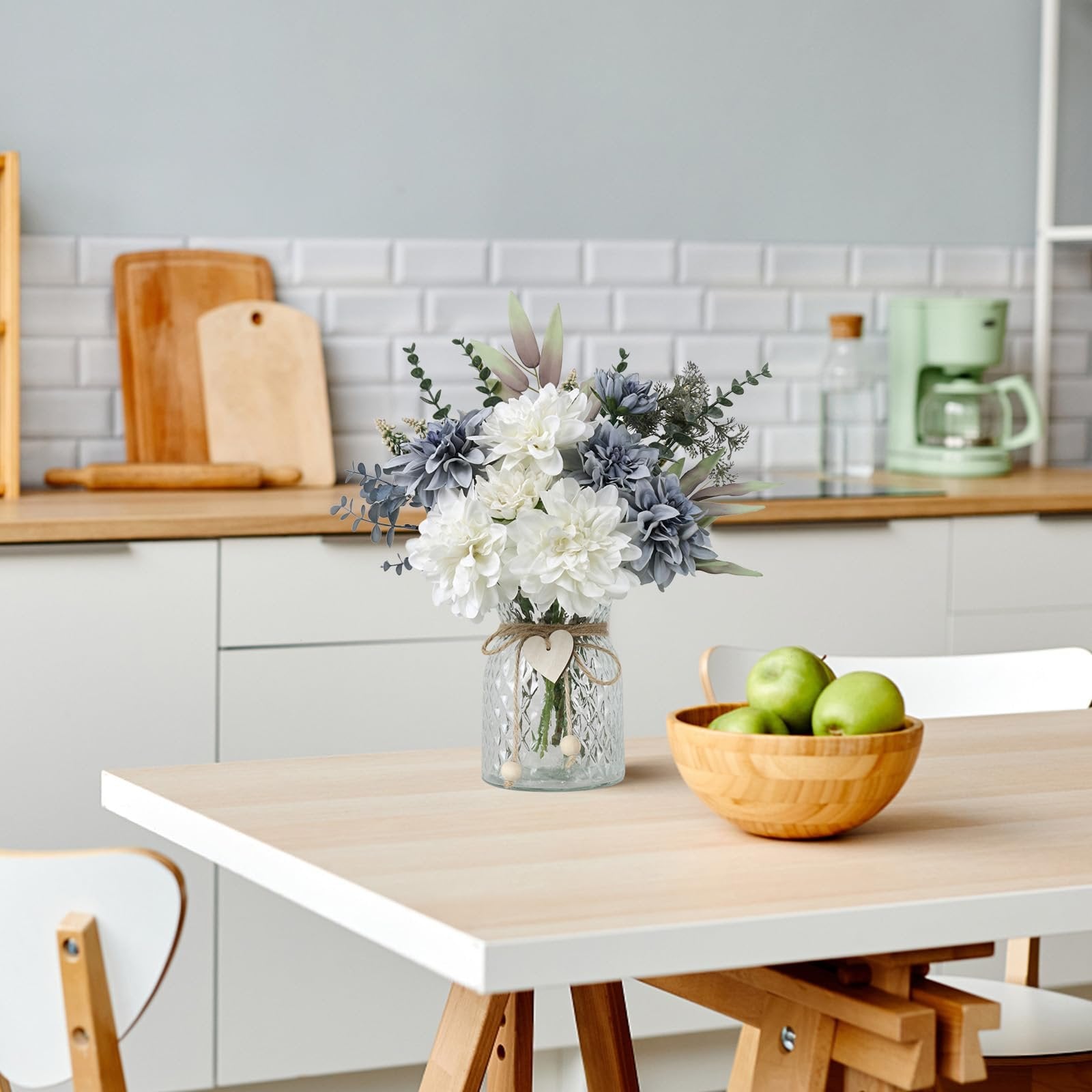 Artificial dahlia flower arrangement in a glass vase on a wooden table with a bowl of green apples